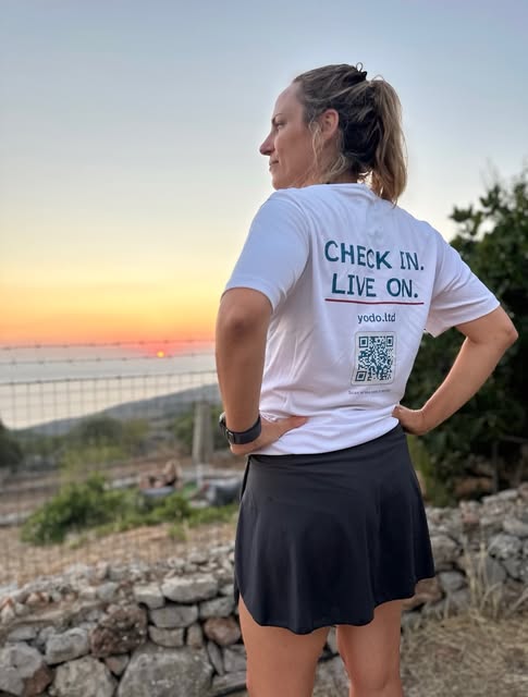Thea Kouraki, founder of Y.O.D.O., photographed from behind at sunset in a white Y.O.D.O. t-shirt reading Check in. Live on., looking out over the sea.